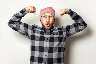 Şapkalı sakallı genç bir adamın portresi, ekoseli bir gömlek izole edilmiş ışık arka planında şaşırmış bir yüze sahip pazıları gösteriyor. Duygusal bir yüz. Gücün hareketi.