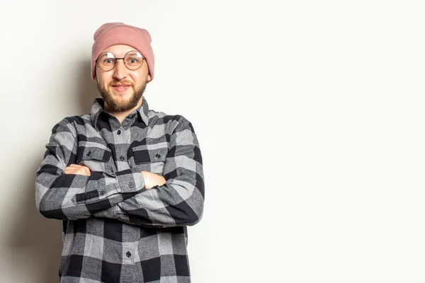 Portrait of a young man with a beard in a hat, a plaid shirt and glasses with arms crossed on his chest on an isolated light background. Emotional face.