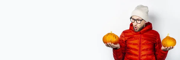赤いジャケットと帽子の若い怖い男は 2つのカボチャを保持し 軽い背景にカボチャを見てください ハロウィン お祝い 秋の概念 バナー — ストック写真