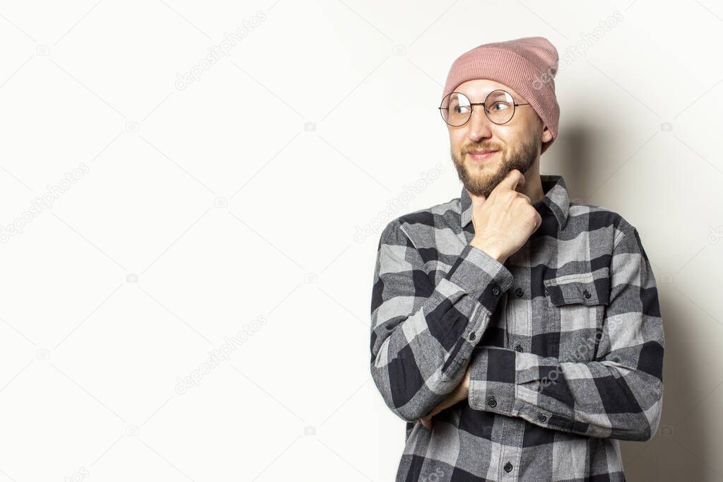 Portrait of a young man with a beard in a hat, a plaid shirt and glasses looks away with an embarrassed face on an isolated light background. Emotional face.