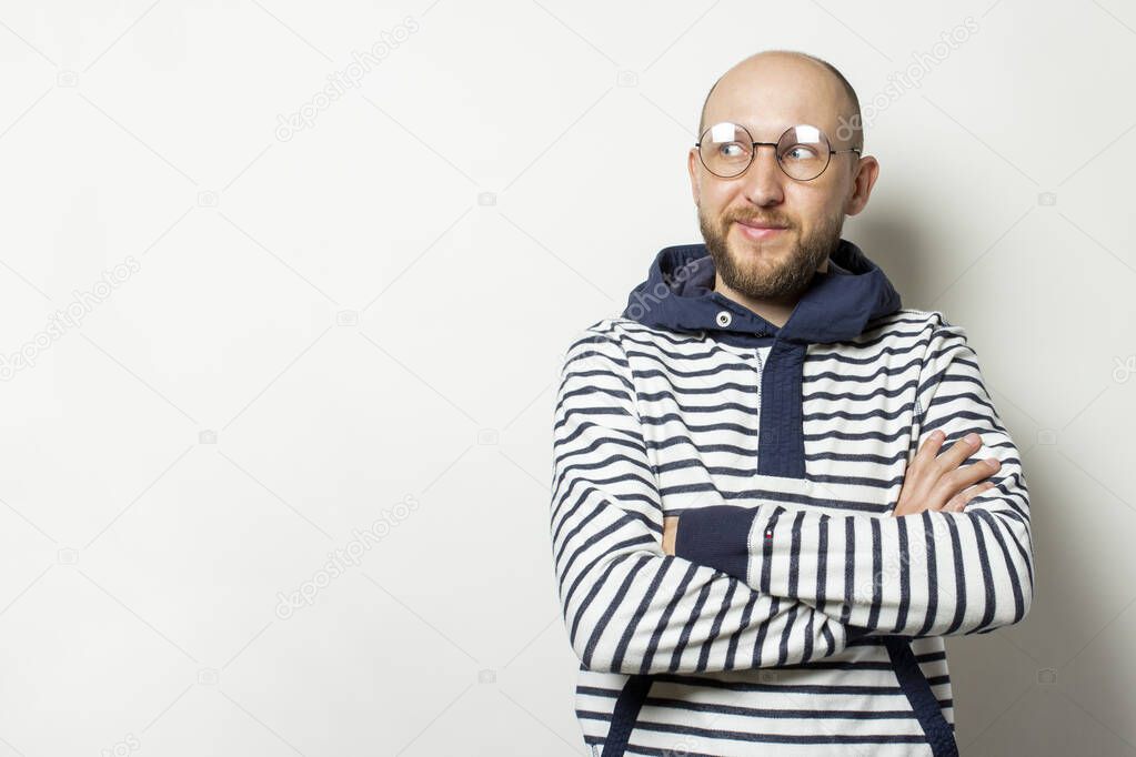 Portrait of a Bald Young man with a beard in glasses of a jacket with a hood looks away on an isolated light background. Emotional face.