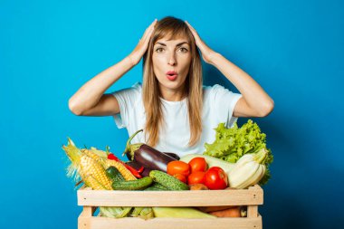 Şaşırmış yüzlü genç bir kadın mavi arka planda taze sebzelerle dolu bir kutu tutuyor. İyi hasat kavramı, doğal ürün.