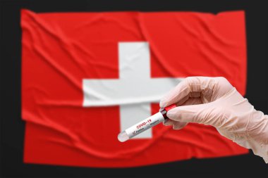 Bir doktor İsviçre bayrağının önünde koronavirüs için pozitif kan testi yapılan bir tüp tutuyor. Covid-19 salgını, enfeksiyon, salgın, zatürree.