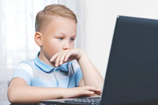 Um menino branco europeu pequeno e sério bonito olha atentamente para a tela do laptop em casa. Estudo moderno ou trabalho em casa conceito. Crianças e gadgets . — Fotografia de Stock