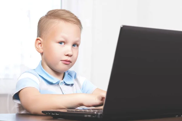 Menino europeu bonito bonito novo com olhos azuis olha para a câmera enquanto sentado e estudando em casa em um laptop. O conceito de educação moderna e trabalho . — Fotografia de Stock