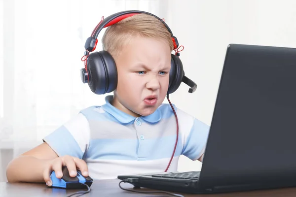 Menino bonito europeu pequeno senta-se em um laptop em fones de ouvido com um microfone com um mouse de computador em sua mão e joga um jogo online. A criança está irritada e insatisfeita com sua perda e cl — Fotografia de Stock