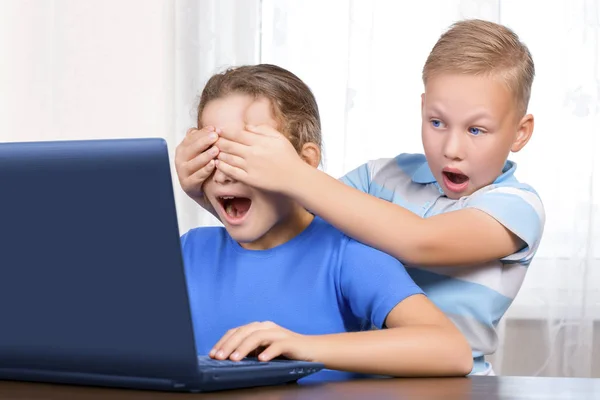 小さなかわいい男の子はノートパソコンのモニターで広い目と口で見て、自分の手で美しい女の子の目を閉じます。子供の安全オンラインコンセプト。インターネット上の禁止コンテンツ. — ストック写真