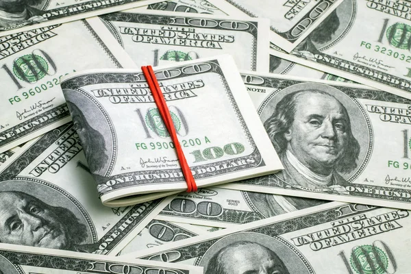 Пучок американських готівок складений навпіл з червоною гумовою стрічкою на грошовому фоні з розкиданих банкнот вартістю в сто доларів. Бізнес, фінанси, торгівля, прибуток, гроші — стокове фото