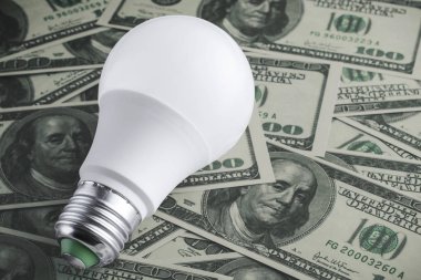 Etrafa saçılmış yüz dolarlık banknotlardan elde edilen para arkaplanındaki enerji tasarrufu ampulünün yakın görüntüsü. Modern teknoloji ve yenilikleri kullanarak para biriktirme kavramı. Anlaşıldı. 