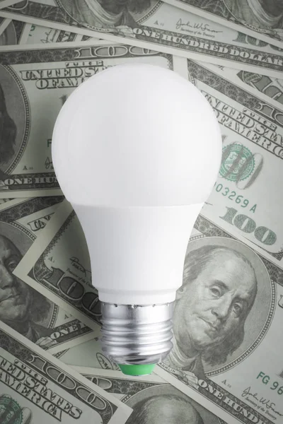 Vue rapprochée d'une lampe à économie d'énergie blanche sur fond d'argent à partir de billets américains éparpillés à cent dollars. Économiser de l'argent en utilisant le concept moderne d'économie d'énergie des ampoules LED — Photo