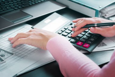 Ofisteki kadın muhasebeci gelir tablosu ve hesap makinesi kullanarak yıllık rapor hazırlıyor. Kar analizi, vergilerin hesaplanması, harcamalar ve ödemeler, tasarruf parası ve bütçe kavramı