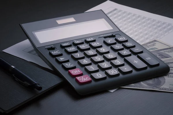 Black accounting calculator, notepad, pen, report with a numerical table and several hundred dollar bills on a dark wooden table. Stock trading, calculation of taxes, profits, expenses and incomes con