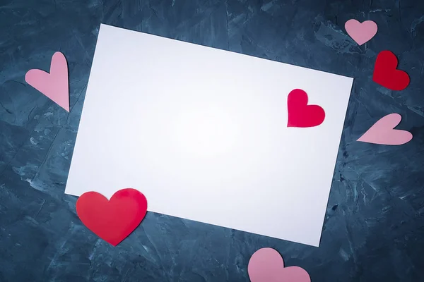 Flat lay tarjeta de felicitación blanca con espacio de copia sobre un fondo texturizado abstracto azul con corazones rojos y rosados dispersos cortados de papel de varias formas . —  Fotos de Stock