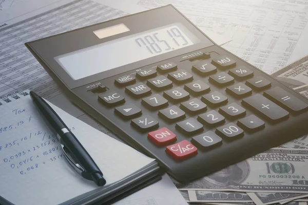 Side view of black accounting calculator, open notebook with notes, pen, documents and reports with tables and several hundred dollar bills. Taxes, profits, payments saving money concept.