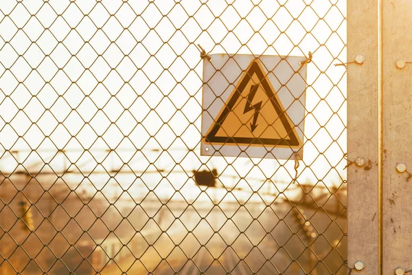 日没時に鉄道線路上の鉄メッシュフェンス上の黄色の高電圧警告標識. — ストック写真