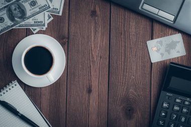 Üzerinde bir fincan kahve, bir kredi kartı, bir hesap makinesi, kalemli bir defter, dolar banknotları ve fotokopi alanı olan bir dizüstü bilgisayar olan ahşap bir kırsal masa..