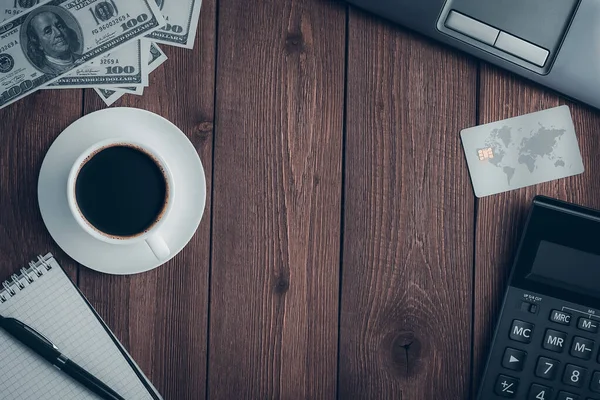 Üzerinde bir fincan kahve, bir kredi kartı, bir hesap makinesi, kalemli bir defter, dolar banknotları ve fotokopi alanı olan bir dizüstü bilgisayar olan ahşap bir kırsal masa..