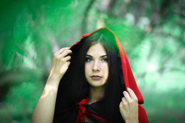Hello summer. Attractive girl with green eyes in a red cloak. Witch among nature and greenery