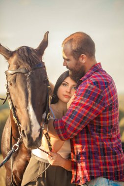 Brunette girl and brutal guy with a beard with a black horse. Sexy couple. Summer trip. Love story clipart