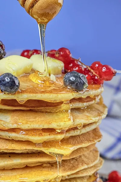 honey from a spoon flows on a stack of pancakes with butter, blueberry and currant