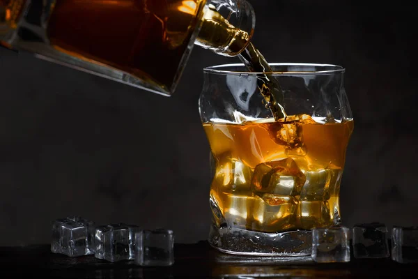 Whiskey bottle áramlik egy pohár jég egy sötét háttér — Stock Fotó