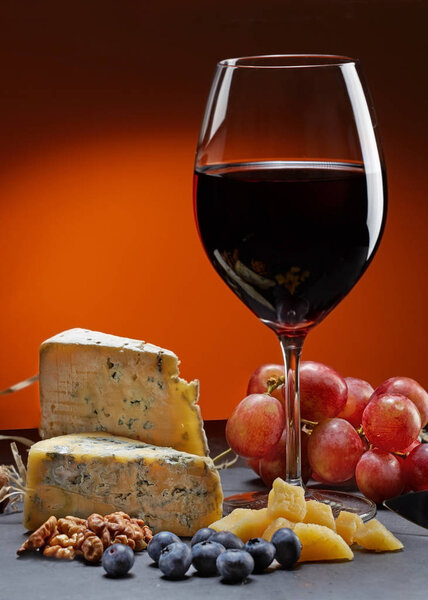 Glass of wine with grapes and a piece of cheese with mold.walnuts, Parmesan cheese and blueberries.  Orange background.