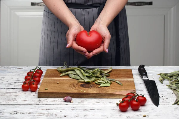 a heart-shaped tomato. a woman\'s hands hold a heart-shaped tomato against a gray black-striped apron on a white kitchen. cooking healthy food. Love and health concept. cook with love.