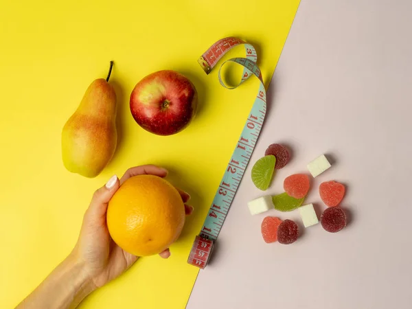 Рука жінки робить вибір на користь правильного харчування. на рожево-жовтому тлі викладаються корисні фрукти, апельсин яблучної груші та шкідливі цукерки. Посередині знаходиться вимірювальна стрічка — стокове фото