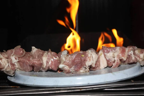 Trozos crudos de cerdo empalados en un pincho acostado en una tapa de metal. Las llamas del fuego detrás — Foto de Stock