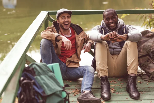 Hombres sonrientes con mochilas sentados en el muelle —  Fotos de Stock