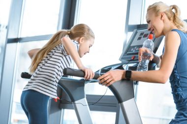Woman trainer looking at small girl workout on treadmill clipart