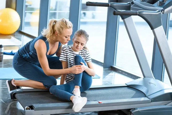 Mujer ayudando niña lesionada rodilla sentado en la cinta de correr —  Fotos de Stock