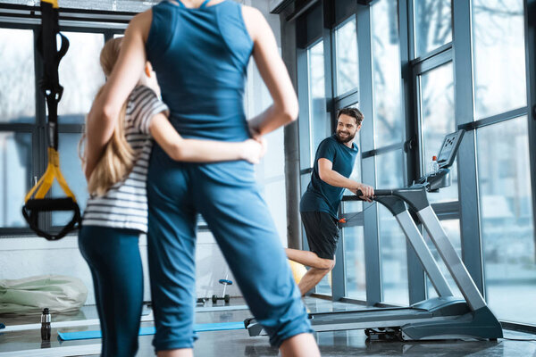 Woman with girl looking at handsome man workout on treadmill at gym