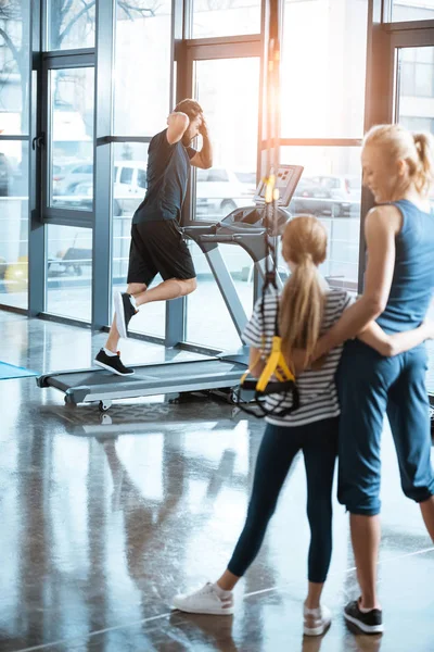 Woman with girl looking at handsome man workout on treadmill at gym — Stock Photo, Image