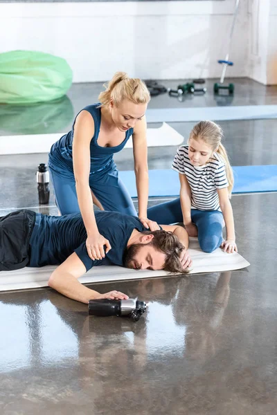 Kvinna med flicka hjälper trött man liggande på matta på gym — Stockfoto