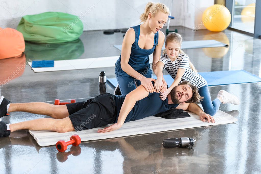 Woman with girl helping tired man lying on mat at gym