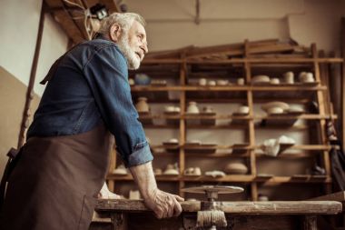 Üst düzey potter ayakta ve raflar atölyesinde seramik ürünler ile karşı masada eğilerek önlük yan görünüm