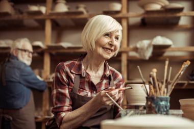 Woman painting clay pot with senior potter at workshop clipart