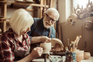 Woman painting clay pot with senior potter at workshop clipart