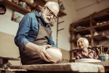Üst düzey potter önlük ve arka planda çalışan kadın ile seramik kase inceleyerek gözlük