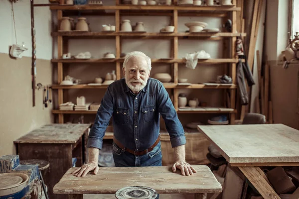 Front view of senior potter standing and leaning on table against shelves with pottery goods at workshop — Stock Photo, Image