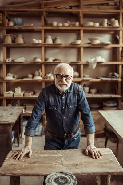 Front view of senior potter standing and leaning on table against shelves with pottery goods at workshop — Stock Photo, Image