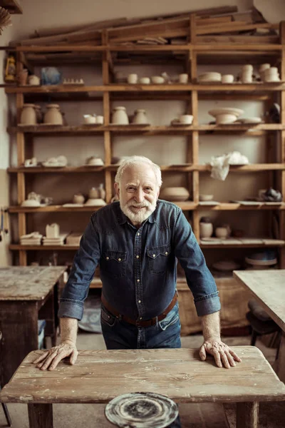 Front view of senior potter standing and leaning on table against shelves with pottery goods at workshop — Stock Photo, Image