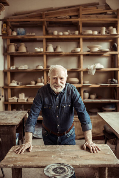 Front view of senior potter standing and leaning on table against shelves with pottery goods at workshop