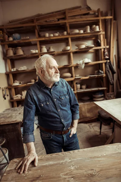 Frontansicht eines älteren Töpfers, der in der Werkstatt auf einem Tisch steht und sich an Regale mit Töpferwaren lehnt — Stockfoto