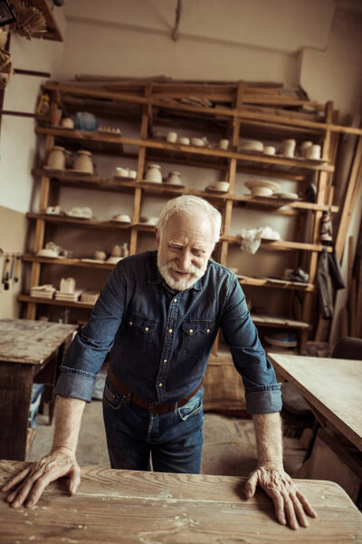 Front view of senior potter standing and leaning on table against shelves with pottery goods at workshop