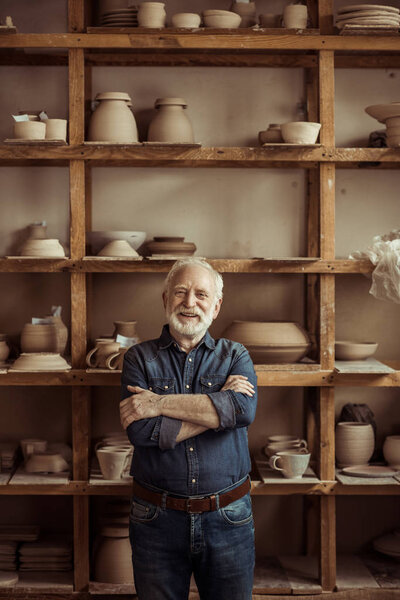 Front view of senior potter standing against shelves with pottery goods at workshop