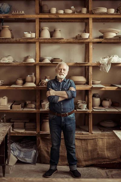 Front view of senior potter standing against shelves with pottery goods at workshop — Stock Photo, Image