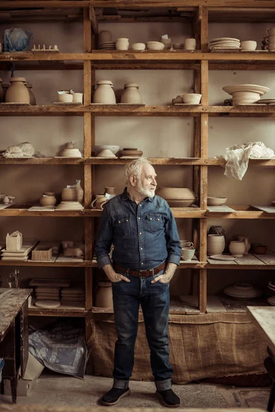 Vooraanzicht van senior potter permanent tegen planken met aardewerk goederen op workshop — Stockfoto