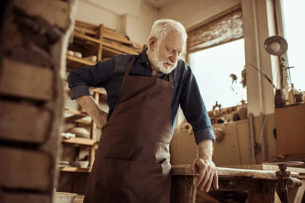 Vooraanzicht van senior potter in schort permanent en leunend op tabel tegen planken met aardewerk goederen op workshop — Stockfoto